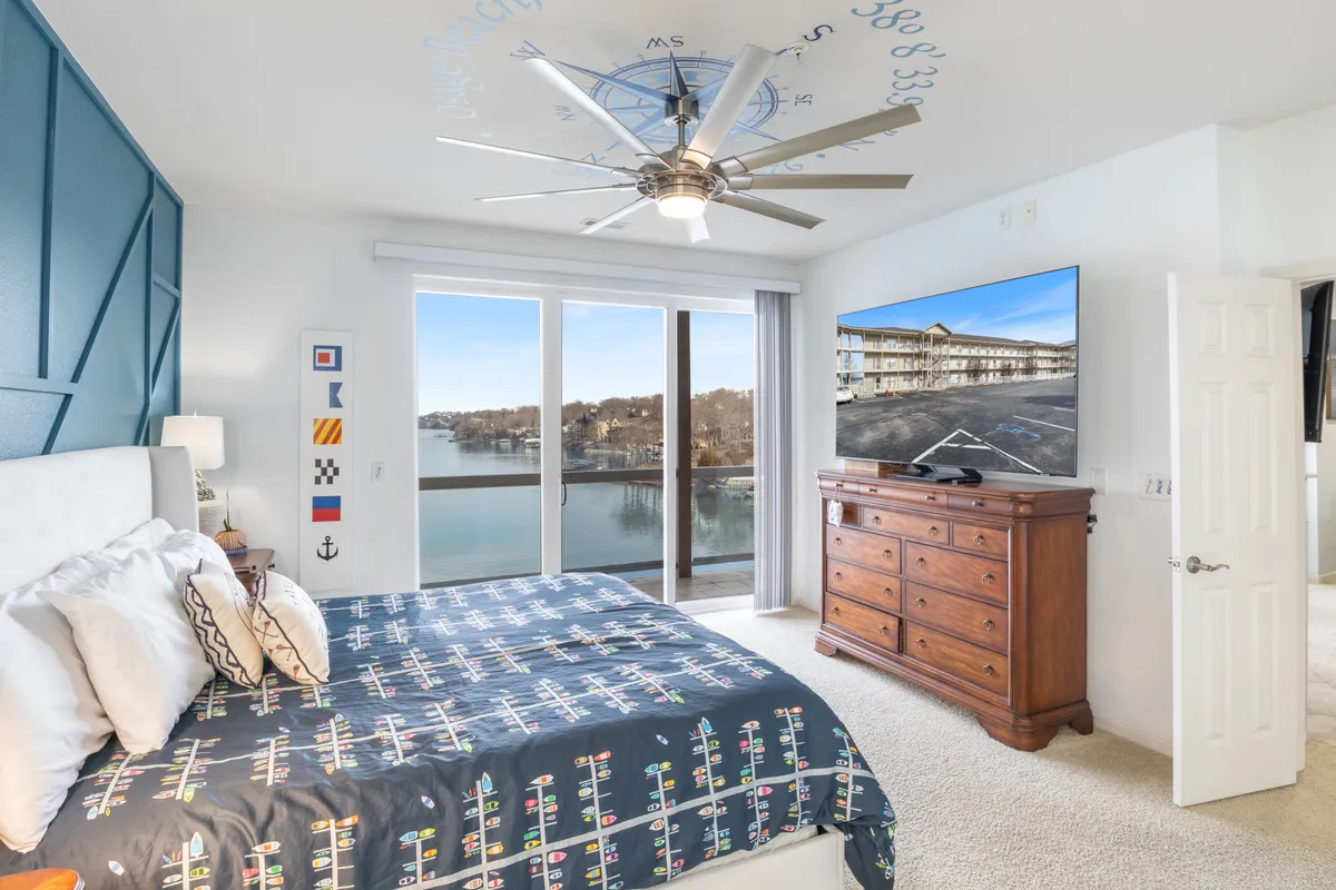 A serene bedroom with a bed made up in crisp luxury linens, decorative pillows, and a large window that offers sweeping views of the lake. The room features a wooden dresser and a television, with calm nautical accents subtly incorporated in the decor. Natural light fills the space, emphasizing its tranquil and rejuvenating atmosphere.