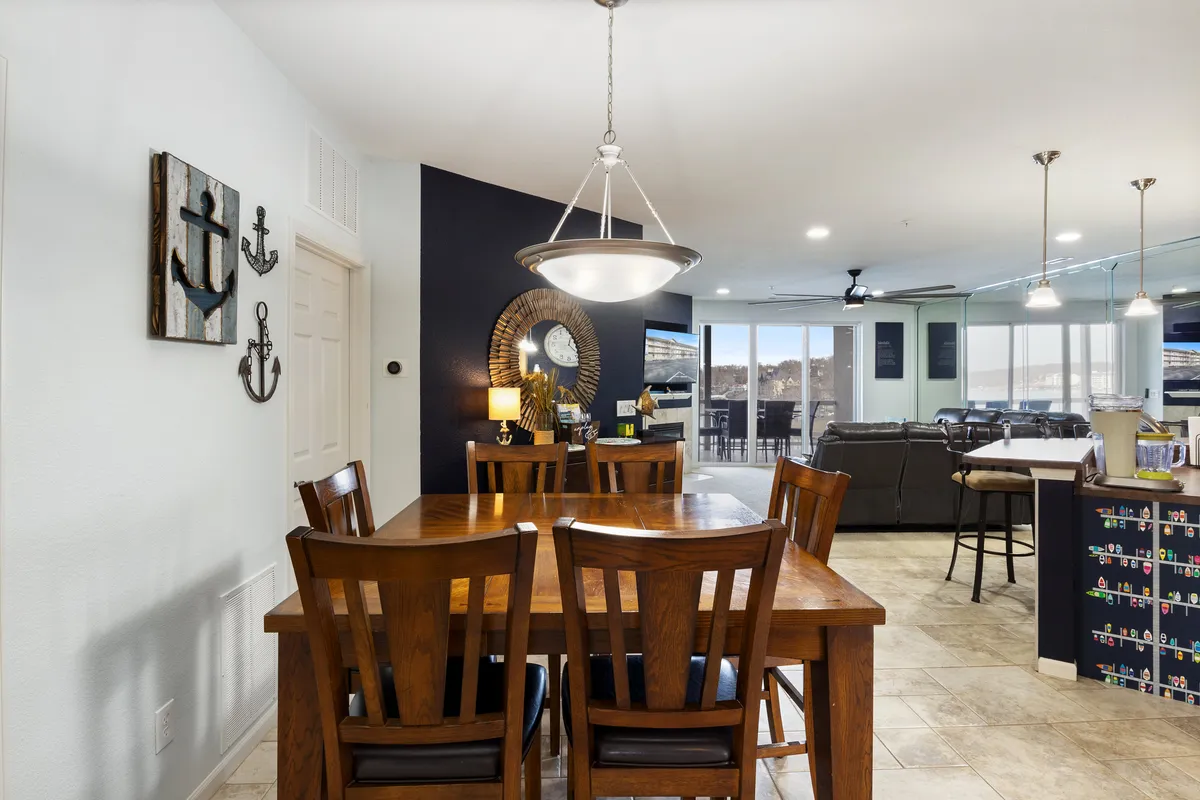 A warm dining area centered around a spacious table with stylish chairs, positioned near large windows that reveal a beautiful lake view. Decorative wall art enhances the space, and the atmosphere is perfect for both intimate family meals and lively group gatherings.