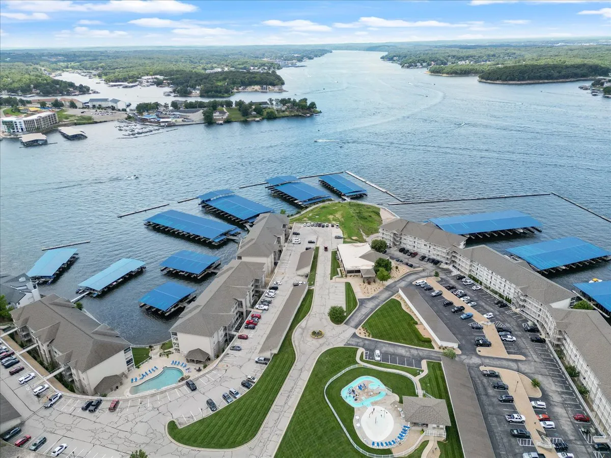 This exterior image presents a sweeping waterfront view with visible boat slips, a landscaped green area, a swimming pool, and residential buildings in the background. It highlights the dock and the direct lake access, with boats docked along the shore.