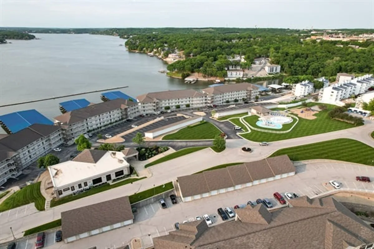 This image captures a scenic lakefront area with views of the lake, a swimming pool, landscaped green spaces, and surrounding residential buildings. The inviting shoreline and open spaces signal opportunities for swimming, picnicking, and outdoor games, all set against the backdrop of Lake of the Ozarks.