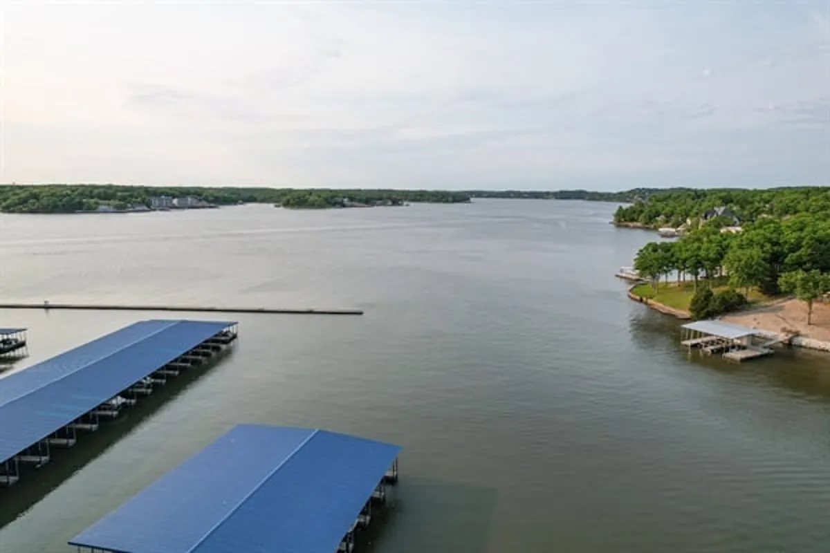 A tranquil lakeside scene shows blue boat docks extending into calm water, surrounded by lush green trees and homes along the shoreline. The docks are clearly visible, emphasizing easy water access for boating, fishing, and water sports. The setting sun bathes the area in warm light, creating a peaceful yet active atmosphere.