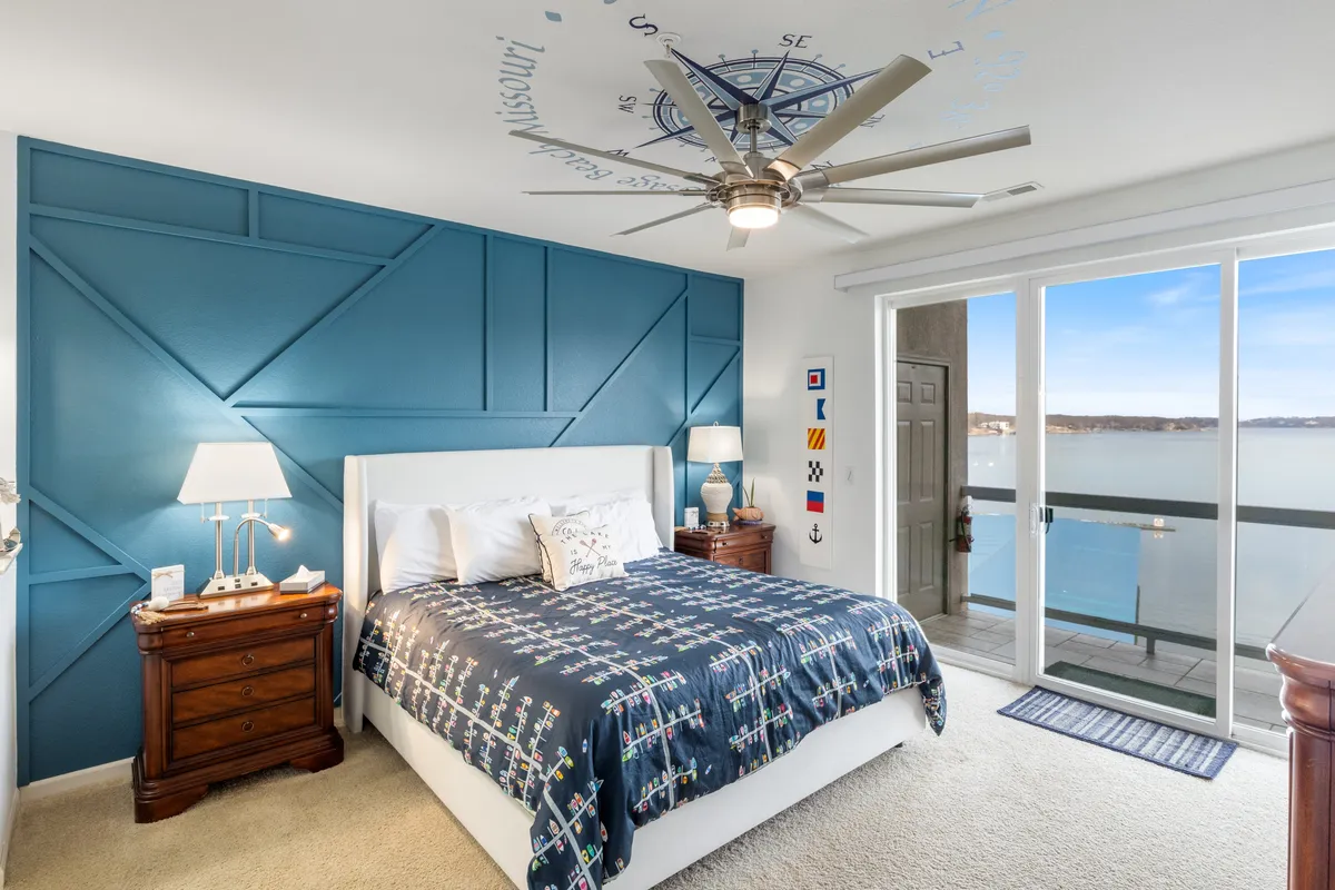 A bedroom featuring a blue accent wall, a bed adorned with nautical-themed bedding, and a sliding glass door that opens onto a balcony. The decor is soothing with a coastal flair, and soft natural light enhances the tranquil mood of the room.