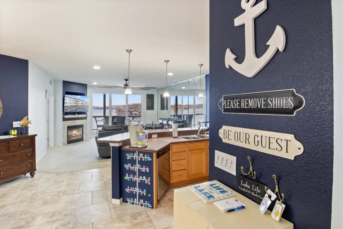 A bright, open kitchen and living area featuring a kitchen island, anchor wall decor, a cozy fireplace, and large windows with beautiful lake views. The space feels inviting and ready for guests to cook, entertain, or simply relax while enjoying the natural light and scenery.