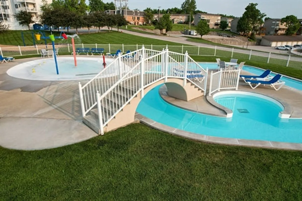 A joyful, sunlit pool area surrounded by lounge chairs, a small bridge over the water, playful water features, and lush green space. Families and groups gather for relaxation and fun, with the lake's natural beauty in the background, alluding to both leisure and discovery.
